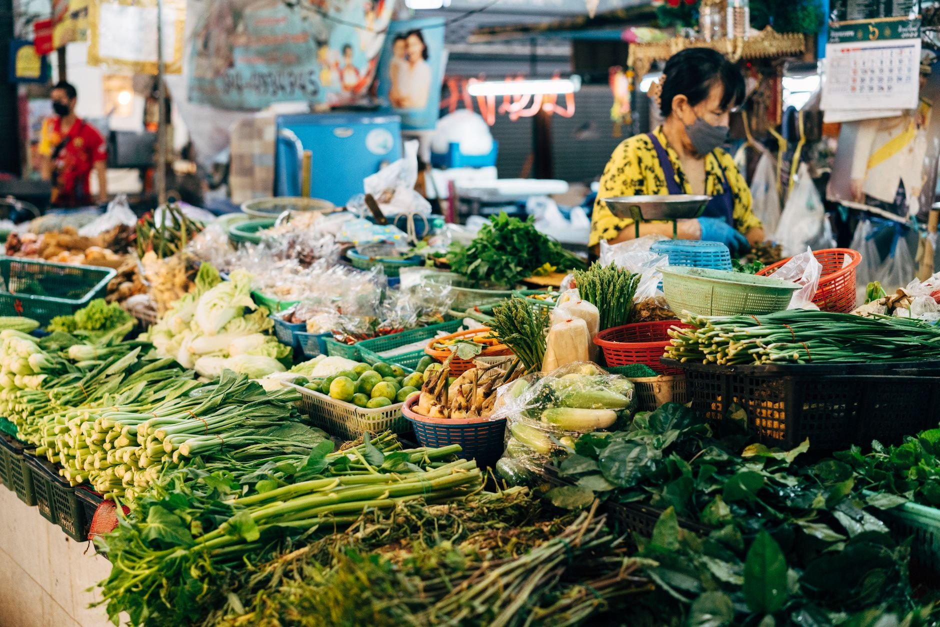 A bustling market stall filled with fresh vegetables and a woman vendor in an urban setting.